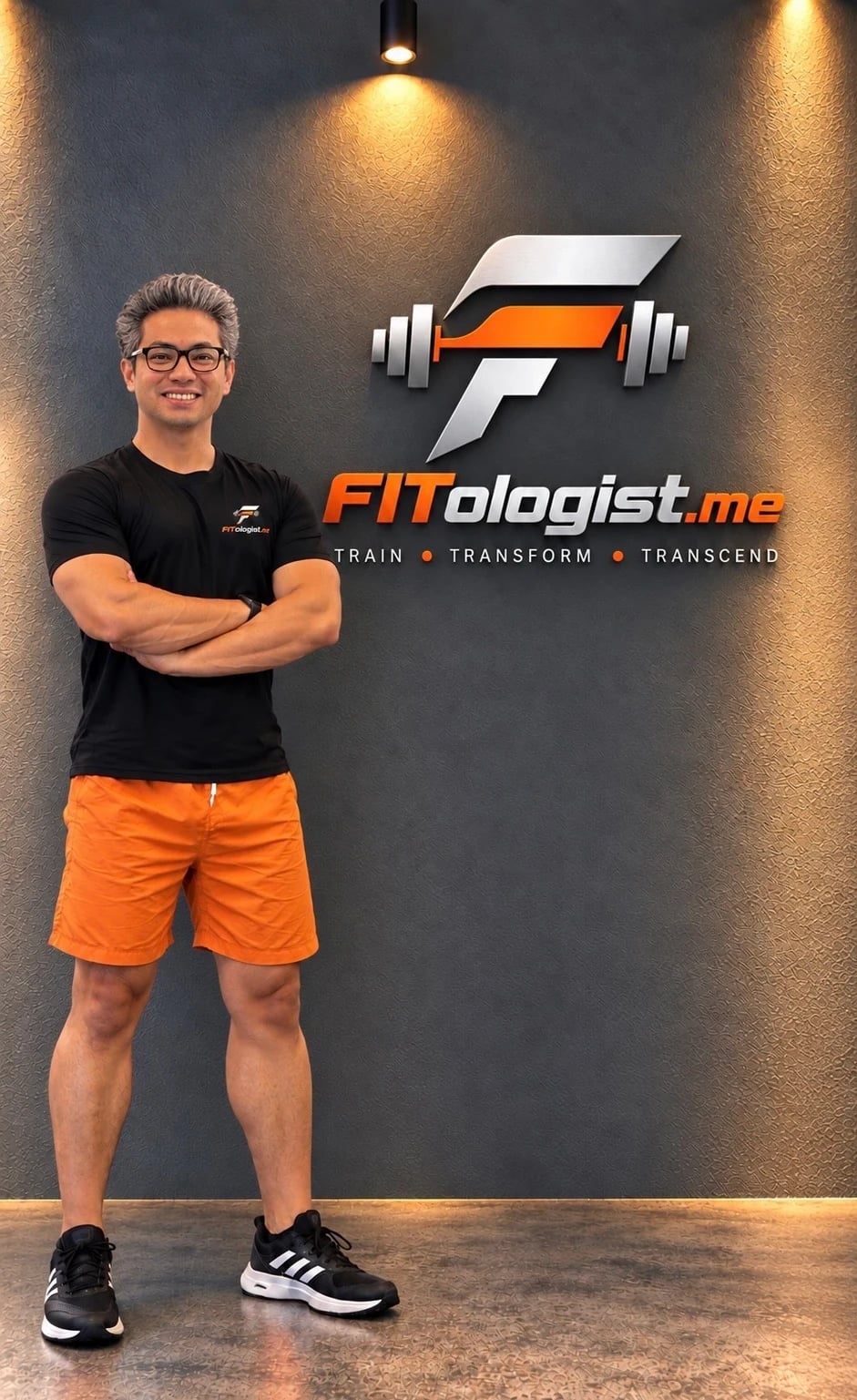 Saeid, full length, arms crossed, standing in front of a textured wall with the FITologist.me sign
